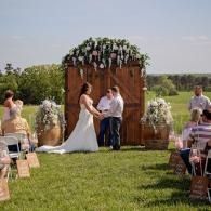 Outdoor wedding ceremony with couple at altar under floral arch, guests seated on grass, officiant nearby