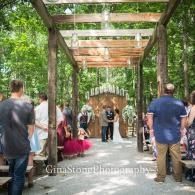 Outdoor wedding ceremony under a wooden pergola with guests seated along a garden aisle