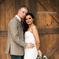 Couple posing in wedding attire against a wooden barn door, holding each other close.