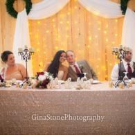 Four people seated at a decorated wedding reception head table with white drapery and warm lights behind them