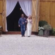 Couple walking arm-in-arm out of a barn doorway, the bride in a white dress and the groom in blue jeans.