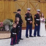 Military wedding party standing on gravel in front of a wooden barn, bride holding bouquet