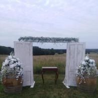 Outdoor wedding arch with white drapes, floral arrangements, and a small table in a grassy field