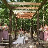 Outdoor wedding ceremony under a wooden pergola in a forest, bride seated at center with bridesmaids in pink dresses.
