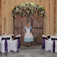 Person standing under a floral arch in a rustic wedding reception hall with decorated tables and chairs