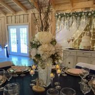 Elegant dining table with white floral centerpiece, candles, glassware, and a tiered cake in a rustic room
