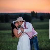 Couple kissing in a grassy field at sunset, with a pink-orange sky behind them