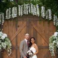 Wedding couple posing before a wooden door with white floral arch and arrangements