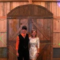 Two people stand smiling in front of large wooden barn doors.