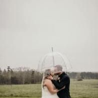 Couple embracing under a clear umbrella in a grassy field under an overcast sky