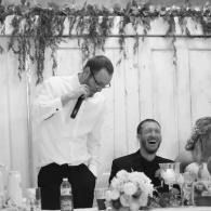 Man in white shirt singing into a microphone at a table, while two guests laugh at a wedding reception.