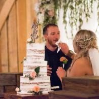 Wedding couple feeding cake beside a tiered white cake with greenery decorations