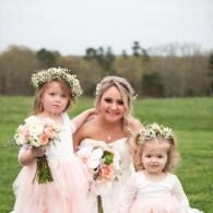 Three people in light pink dresses and flower crowns posing in a grassy field, holding bouquets.