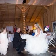 Bride sitting on a man's knee at a wedding reception in a wooden hall, with children nearby