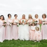 Wedding party posing outdoors in blush and white dresses, holding bouquets on grass.