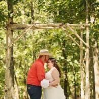 Couple in white and red embracing under a wooden arch in a sunlit forest