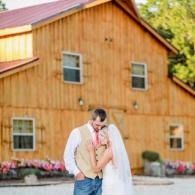 Couple embracing in front of a wooden barn with pink flowers and warm sunlight