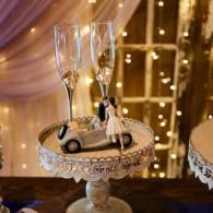 Decorative cake with miniature car and couple topper, set on a lace stand with champagne flutes