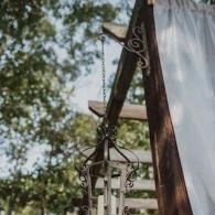 White drapes and a hanging lantern on a dark wooden outdoor pergola with leafy trees behind