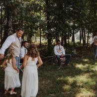 People in white outfits standing and sitting in a shaded wooded area during an outdoor gathering