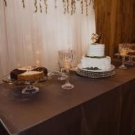 Dessert table with a two-tier white cake, pie, and crystal glasses on a brown tablecloth