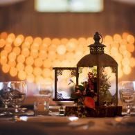 A lantern centerpiece with flowers on a softly lit dinner table, against warm blurred lights.