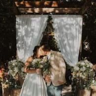 Couple embracing under a floral wedding arch, surrounded by white drapes and greenery.