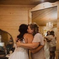 Bride and woman embrace in a rustic wooden room, with a mirror reflecting the scene.