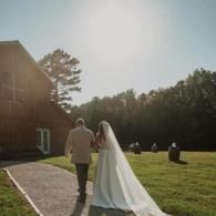 Bride and groom walking hand in hand toward a barn at sunset, with guests seated on the lawn.
