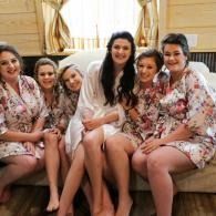 Five women in floral robes sit together on a couch, smiling in a bright room.