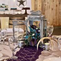 Decorated banquet table with lantern centerpiece, purple runner, and glassware in a rustic room