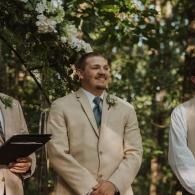 Man in a beige suit smiling at an outdoor ceremony among trees