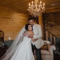 Bride in a white gown and groom in a light jacket embrace in a wood-paneled room under a chandelier.