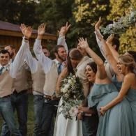 Wedding party cheering outdoors with raised hands, bride holding bouquet, bridesmaids in blue dresses and men in vests