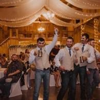 Three men in white shirts and blue ties dance at a warmly lit indoor celebration.