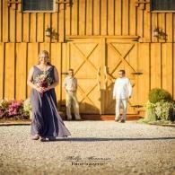 Woman in a dark purple dress holding flowers, walking ahead of two people by a yellow barn door.