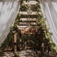 Rustic outdoor wedding altar draped in white fabric and green garlands under a wooden archway