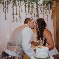 Couple sharing a kiss beside a wedding cake at a reception under hanging greenery
