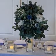 Round floral centerpiece with white and blue flowers on a decorated dining table with candles.