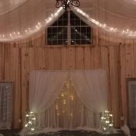 Wooden barn interior with draped white fabric, warm string lights, and small glowing trees by a curtained archway