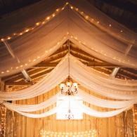 Warmly lit draped ceiling with string lights and a central chandelier in an elegant tented venue