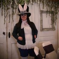 Woman in a black jacket and bunny-ear hat posing indoors beside a chair and white doors.