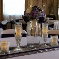 Elegant banquet table with white cloth, candles, and a purple floral centerpiece in a warm-lit room