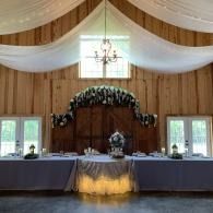 Decorated wedding reception table beneath draped white fabric and floral arch in a barn venue