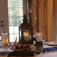 Decorative lantern on a dining table by a sunlit window, with antlers and glassware nearby