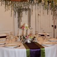 Elegant banquet table with white linens, purple and green runner, and hanging greenery backdrop
