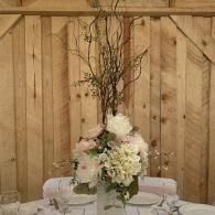 Floral centerpiece with white and blush flowers on a table in front of a wooden barn wall