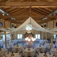 Elegant rustic wedding reception in a barn with draped white fabric, chandeliers, and round tables set with white linens