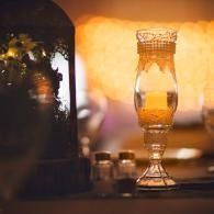 Warm-lit glass candle holder on a table with blurred bottles in the background