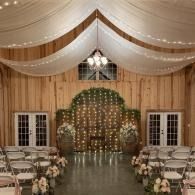 Indoor wedding ceremony setup with draped white fabric, twinkle lights, and rows of chairs facing an arch backdrop